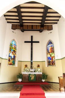 An indoor setting with a central altar decorated with white flowers and candles. Two colorful stained glass windows flank the altar, depicting religious scenes. A large black cross is mounted on the wall above the altar. The ceiling features dark wooden beams, and a red carpet leads up to the altar.