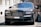 A luxury car with a sleek and polished metallic finish is parked on a cobblestone street. Its grille and headlamps are prominently visible, showcasing an opulent design typical of high-end vehicles.