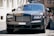 A luxury car with a sleek and polished metallic finish is parked on a cobblestone street. Its grille and headlamps are prominently visible, showcasing an opulent design typical of high-end vehicles.
