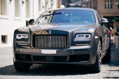 A luxury car with a sleek and polished metallic finish is parked on a cobblestone street. Its grille and headlamps are prominently visible, showcasing an opulent design typical of high-end vehicles.
