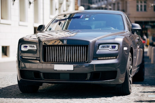 A luxury car with a sleek and polished metallic finish is parked on a cobblestone street. Its grille and headlamps are prominently visible, showcasing an opulent design typical of high-end vehicles.