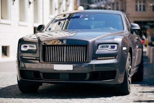 A luxury car with a sleek and polished metallic finish is parked on a cobblestone street. Its grille and headlamps are prominently visible, showcasing an opulent design typical of high-end vehicles.
