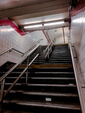 A dimly lit underground staircase with metal handrails and white tiled walls, featuring a red-tiled border near the ceiling. Fluorescent lights illuminate the upper part of the stairwell, and the stairs appear slightly worn.