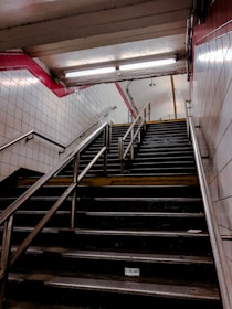A dimly lit underground staircase with metal handrails and white tiled walls, featuring a red-tiled border near the ceiling. Fluorescent lights illuminate the upper part of the stairwell, and the stairs appear slightly worn.
