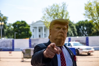 A person wearing a Donald Trump mask and a suit is pointing towards the camera with a background that includes the White House and surrounding barricades. Trees and a parked police car are also visible.