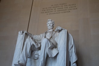 A large, seated statue of a man is crafted from white marble, set against a beige stone wall with an engraved inscription honoring his memory.
