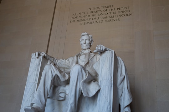 A large, seated statue of a man is crafted from white marble, set against a beige stone wall with an engraved inscription honoring his memory.