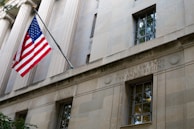 A stone building facade with columns and a prominent window. A United States flag is displayed on a flagpole extending from the building. Engraved in the stone wall is the phrase 'WHERE LAW ENDS TYRANNY BEGINS'. The architectural style suggests a governmental or institutional building.