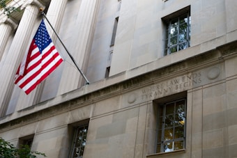 A stone building facade with columns and a prominent window. A United States flag is displayed on a flagpole extending from the building. Engraved in the stone wall is the phrase 'WHERE LAW ENDS TYRANNY BEGINS'. The architectural style suggests a governmental or institutional building.