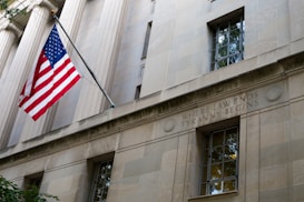 A stone building facade with columns and a prominent window. A United States flag is displayed on a flagpole extending from the building. Engraved in the stone wall is the phrase 'WHERE LAW ENDS TYRANNY BEGINS'. The architectural style suggests a governmental or institutional building.