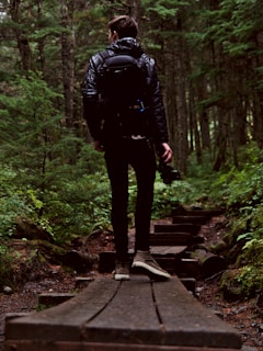 A person in outdoor attire is walking along a wooden path surrounded by dense forest. The individual is carrying a backpack and holding a camera. The scene is lush with green foliage and towering trees.