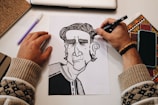 A person's hands are in the process of drawing a black and white cartoon-like character on paper. The workspace includes a laptop, a notebook, a pen, a phone, and a colored decorative item. The person is wearing a bracelet and a sweater with a pattern.