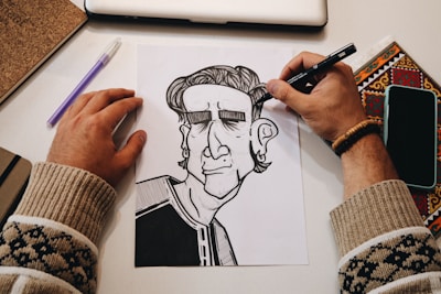 A person's hands are in the process of drawing a black and white cartoon-like character on paper. The workspace includes a laptop, a notebook, a pen, a phone, and a colored decorative item. The person is wearing a bracelet and a sweater with a pattern.