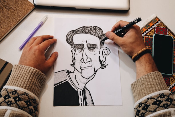 A person's hands are in the process of drawing a black and white cartoon-like character on paper. The workspace includes a laptop, a notebook, a pen, a phone, and a colored decorative item. The person is wearing a bracelet and a sweater with a pattern.