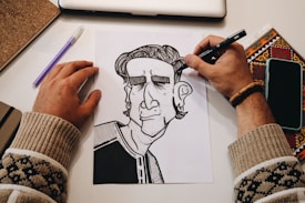 A person's hands are in the process of drawing a black and white cartoon-like character on paper. The workspace includes a laptop, a notebook, a pen, a phone, and a colored decorative item. The person is wearing a bracelet and a sweater with a pattern.