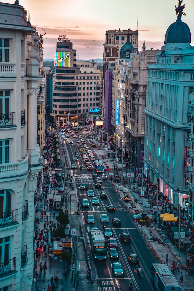 An elevated dusk view down Gran Vía, traffic and crowds streaming between illuminated buildings toward the skyline.