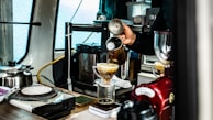 A person is making coffee using a pour-over method. Coffee is being poured from a kettle into a cone-shaped dripper, placed on top of a glass container. The setting includes various coffee-making equipment, such as a scale, grinder, and kettle, suggesting a professional or dedicated homemade setup. The scene is indoors with a mixture of metallic and wooden surfaces.