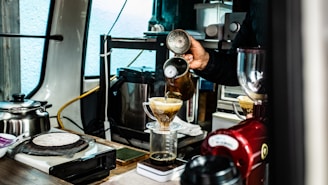 A person is making coffee using a pour-over method. Coffee is being poured from a kettle into a cone-shaped dripper, placed on top of a glass container. The setting includes various coffee-making equipment, such as a scale, grinder, and kettle, suggesting a professional or dedicated homemade setup. The scene is indoors with a mixture of metallic and wooden surfaces.