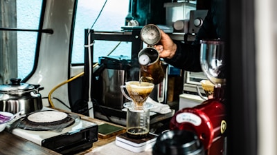 A person is making coffee using a pour-over method. Coffee is being poured from a kettle into a cone-shaped dripper, placed on top of a glass container. The setting includes various coffee-making equipment, such as a scale, grinder, and kettle, suggesting a professional or dedicated homemade setup. The scene is indoors with a mixture of metallic and wooden surfaces.