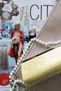 A fashion magazine layout featuring accessories and clothing items. A model walks down the street in a red trench coat and brown outfit, accompanied by text about New York Fashion Week. Various jewelry pieces, including statement necklaces and earrings, surround the magazine page. Prominent fashion elements like a snakeskin clutch, a gold glittery purse, and a pearl necklace add texture and shine.