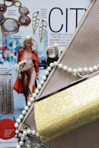 A fashion magazine layout featuring accessories and clothing items. A model walks down the street in a red trench coat and brown outfit, accompanied by text about New York Fashion Week. Various jewelry pieces, including statement necklaces and earrings, surround the magazine page. Prominent fashion elements like a snakeskin clutch, a gold glittery purse, and a pearl necklace add texture and shine.