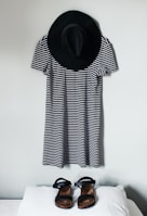 A black and white striped dress is hung on a wall with a black wide-brimmed hat above it. In front of the dress, a pair of black sandals is placed on a white surface.