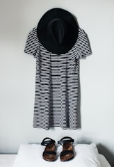 A black and white striped dress is hung on a wall with a black wide-brimmed hat above it. In front of the dress, a pair of black sandals is placed on a white surface.