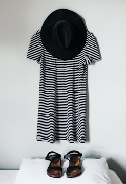 A black and white striped dress is hung on a wall with a black wide-brimmed hat above it. In front of the dress, a pair of black sandals is placed on a white surface.