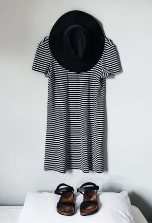 A black and white striped dress is hung on a wall with a black wide-brimmed hat above it. In front of the dress, a pair of black sandals is placed on a white surface.