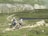Two cyclists are navigating a rugged mountain trail. The landscape features lush green hills, a rocky path, and a small body of water in the distance. Both cyclists are wearing helmets and backpacks, emphasizing the outdoor adventure theme.
