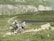 Two cyclists are navigating a rugged mountain trail. The landscape features lush green hills, a rocky path, and a small body of water in the distance. Both cyclists are wearing helmets and backpacks, emphasizing the outdoor adventure theme.