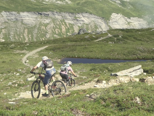 Two cyclists are navigating a rugged mountain trail. The landscape features lush green hills, a rocky path, and a small body of water in the distance. Both cyclists are wearing helmets and backpacks, emphasizing the outdoor adventure theme.