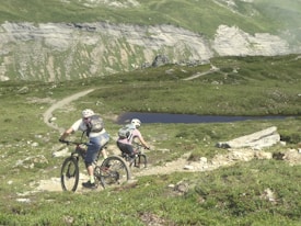 Two cyclists are navigating a rugged mountain trail. The landscape features lush green hills, a rocky path, and a small body of water in the distance. Both cyclists are wearing helmets and backpacks, emphasizing the outdoor adventure theme.