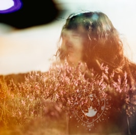 A dreamy, blurred portrait of a person amidst a field of pink flowers. The image has light leaks or an overlay, creating an ethereal effect.