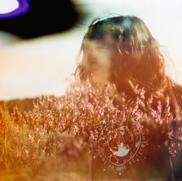 A dreamy, blurred portrait of a person amidst a field of pink flowers. The image has light leaks or an overlay, creating an ethereal effect.