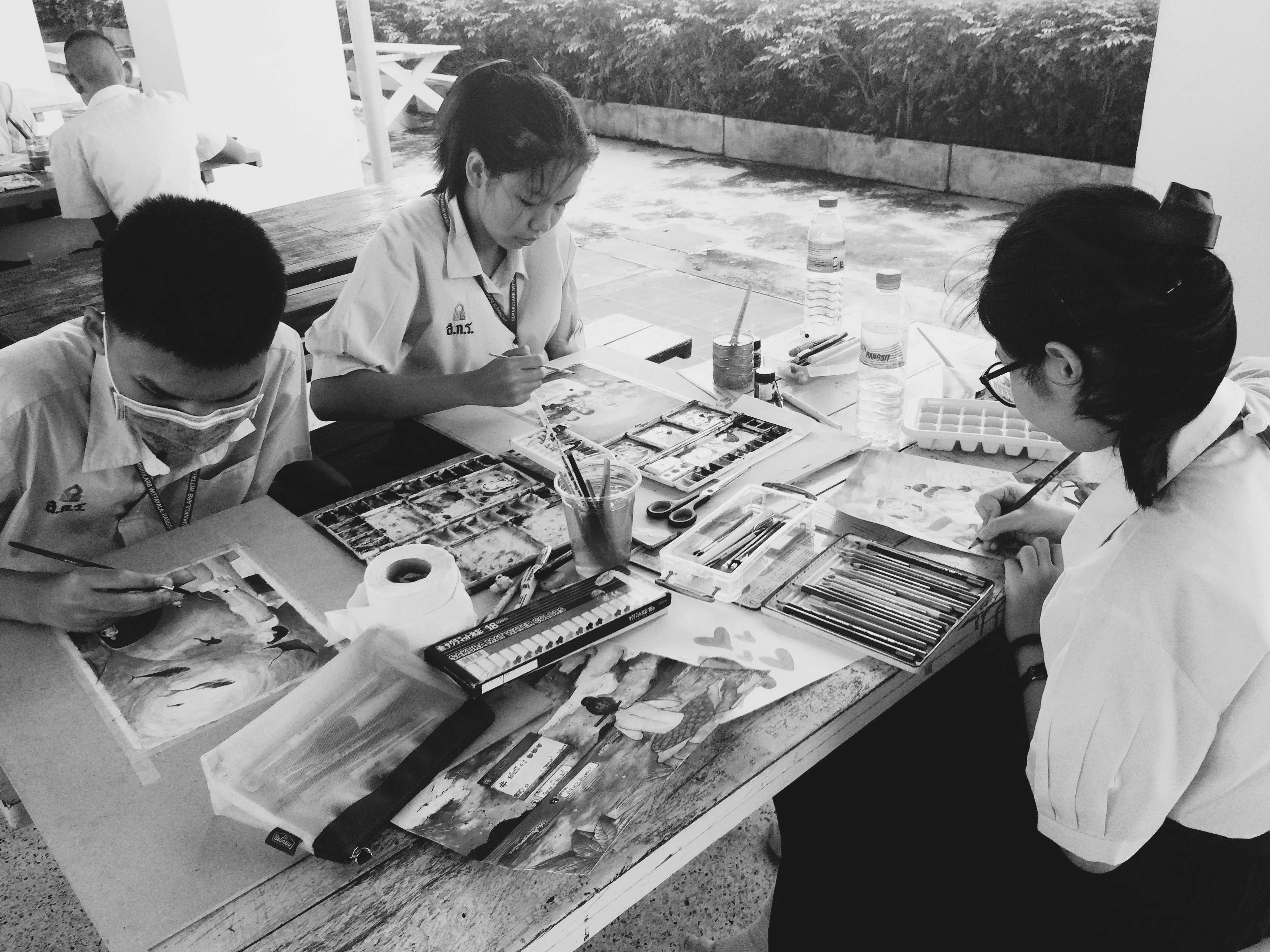 Young students painting at easels outdoors wearing colorful aprons, focused on their individual canvas