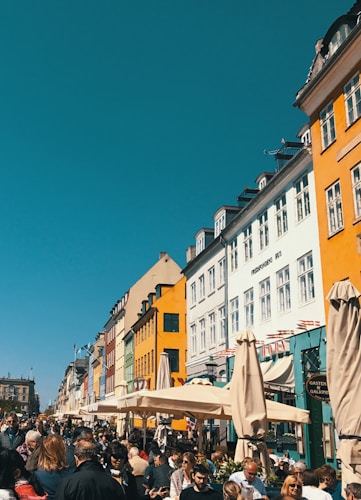 A lively street scene featuring a bustling crowd of people under a clear blue sky. The street is lined with colorful buildings, each with a unique shade ranging from orange to blue, creating a vibrant urban landscape. Outdoor seating with umbrellas suggests a caf&eacute; or restaurant setting, adding to the lively atmosphere.