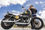 A man stands beside a Harley-Davidson motorcycle under a clear blue sky. The motorcycle is detailed with custom artwork on the fuel tank and features a shiny chrome exhaust. The man wears a cap and a Harley-Davidson jacket, adding to the biker aesthetic.