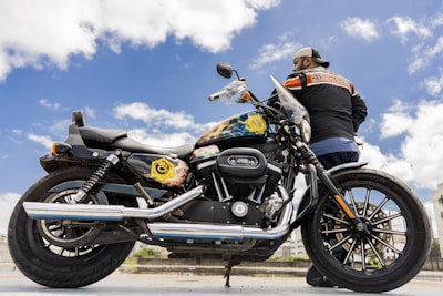 A man stands beside a Harley-Davidson motorcycle under a clear blue sky. The motorcycle is detailed with custom artwork on the fuel tank and features a shiny chrome exhaust. The man wears a cap and a Harley-Davidson jacket, adding to the biker aesthetic.