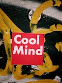 A vibrant red square with the words 'Cool Mind' in bold white lettering is centered on a textured surface. The background is an abstract mix of black, white, and thick yellow brushstrokes. The colors and style create a striking contrast.