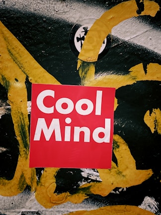 A vibrant red square with the words 'Cool Mind' in bold white lettering is centered on a textured surface. The background is an abstract mix of black, white, and thick yellow brushstrokes. The colors and style create a striking contrast.