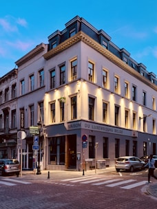 A European street corner with a three-story building that has 'Maison du Luxembourg Belge' written on it. The architecture features classic design elements with large windows and decorative moldings. The scene is set during the evening with streetlights casting a warm glow. There are several road signs and parked cars nearby, and a person is crossing the street.