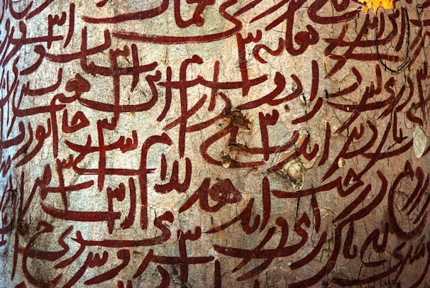 Intricate Arabic calligraphy in deep red is etched onto a textured stone surface. The script is decorative and flowing, creating a sense of artistic expression and cultural heritage.
