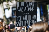 A protest sign with white text on a black background reads, 'If the climate was a bank you would have saved it already.' The sign is held above a crowd of people, likely participating in a climate change demonstration.