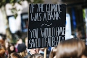 A protest sign with white text on a black background reads, 'If the climate was a bank you would have saved it already.' The sign is held above a crowd of people, likely participating in a climate change demonstration.