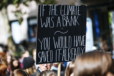 A protest sign with white text on a black background reads, 'If the climate was a bank you would have saved it already.' The sign is held above a crowd of people, likely participating in a climate change demonstration.