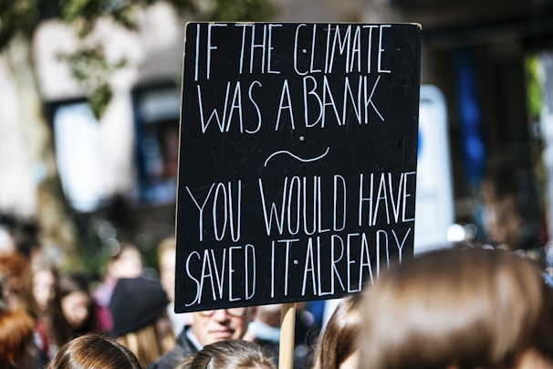 A protest sign with white text on a black background reads, 'If the climate was a bank you would have saved it already.' The sign is held above a crowd of people, likely participating in a climate change demonstration.