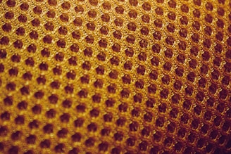 A close-up view of a textured surface featuring a pattern of interconnected hexagonal shapes, resembling a honeycomb. The material appears to be woven, with a warm golden hue.