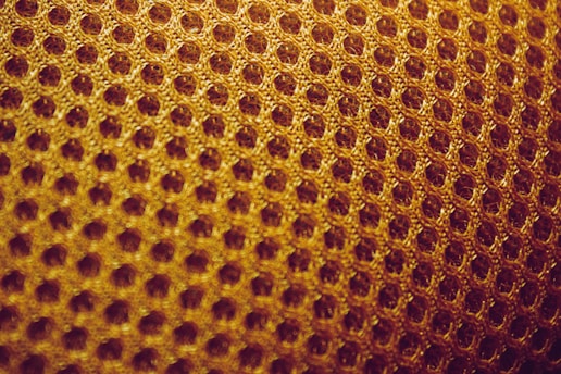 A close-up view of a textured surface featuring a pattern of interconnected hexagonal shapes, resembling a honeycomb. The material appears to be woven, with a warm golden hue.