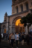 A large crowd gathers in front of a grand cathedral featuring detailed arched entrances lit by warm lighting. The architecture includes intricate stone carvings and tall columns. People are casually dressed, some holding drinks, indicating a social event or gathering.